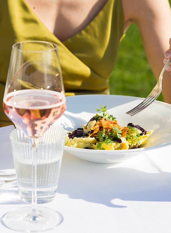 Dettaglio di un piatto in tavola, calice di vino e donna seduta.