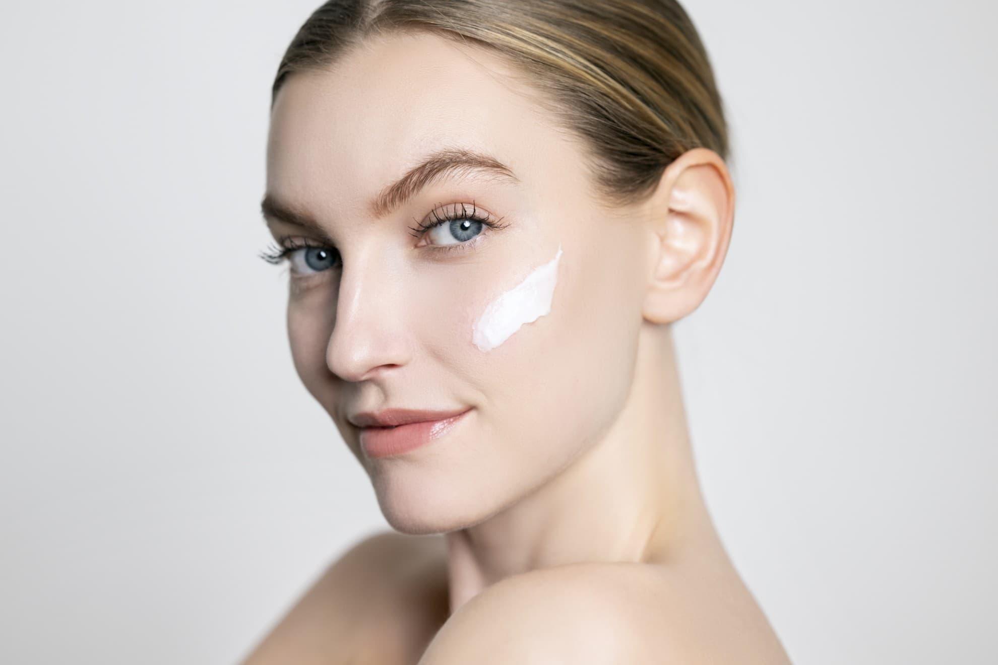 Viso di donna con crema applicata sulla guancia. Viso pulito, pelle luminosa, primo piano su sfondo neutro.