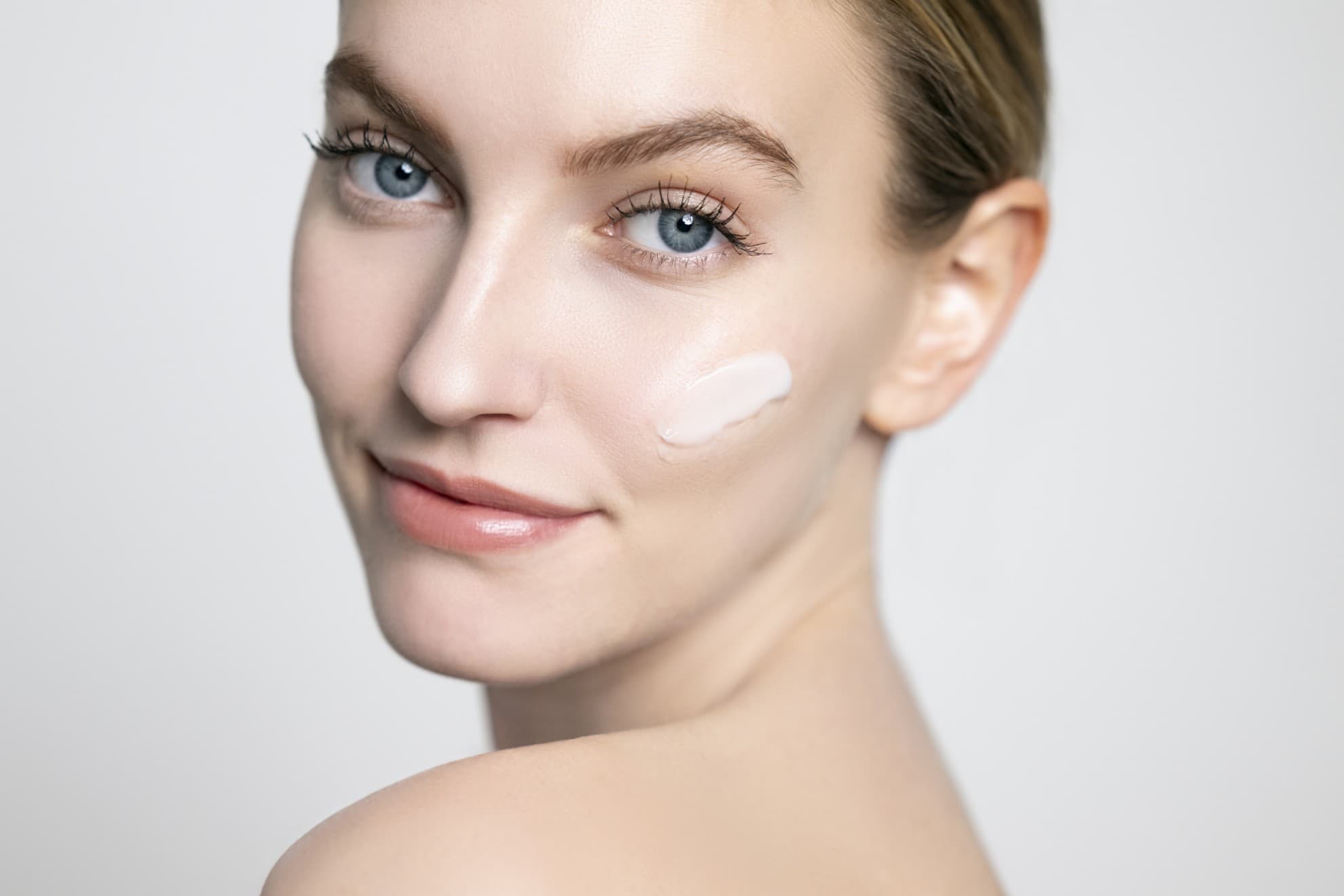 Viso di donna con crema applicata sulla guancia. Viso pulito, pelle luminosa, primo piano su sfondo neutro.