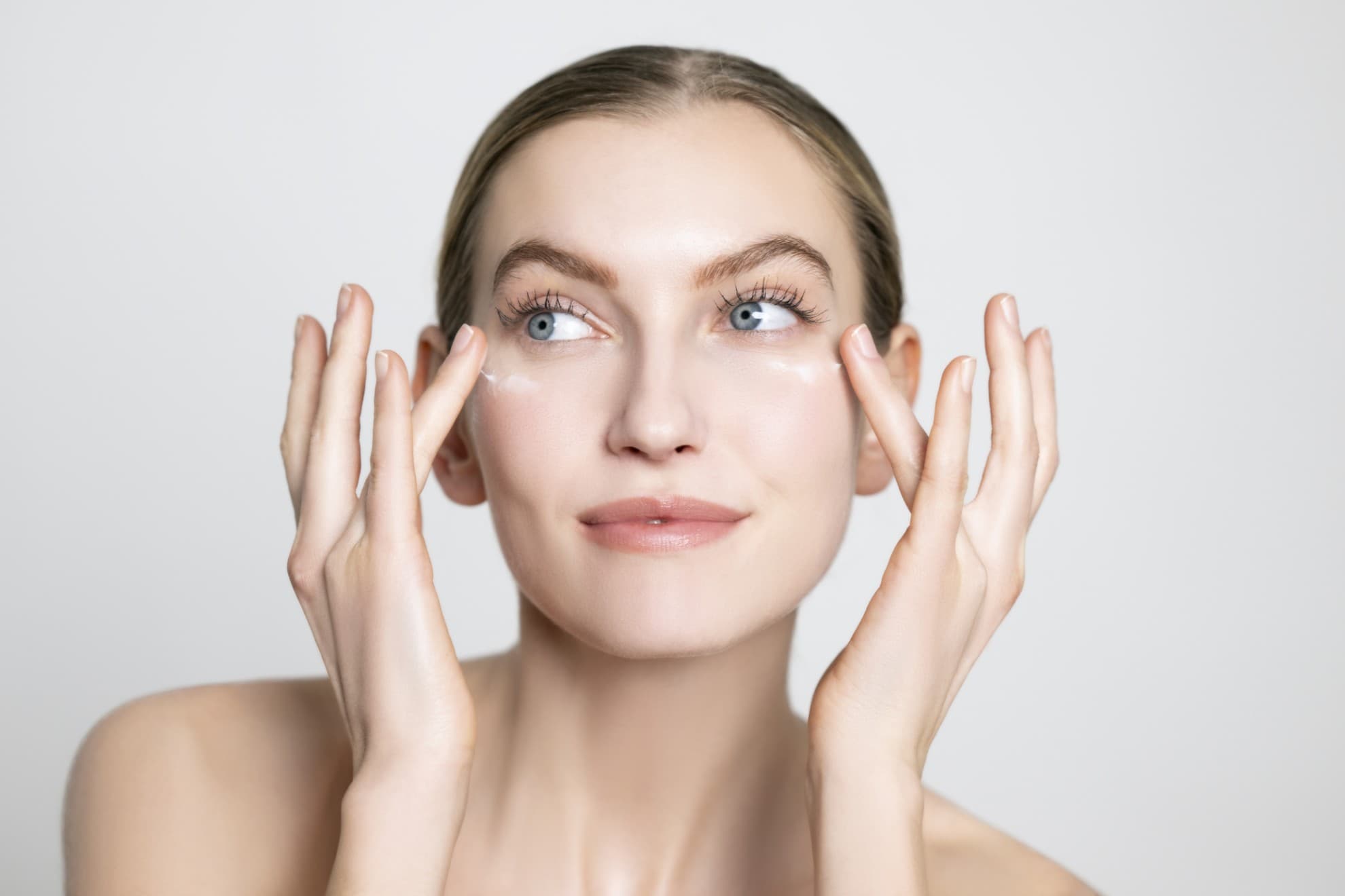 Donna applica crema contorno occhi con le dita, viso pulito, pelle luminosa, primo piano su sfondo neutro.