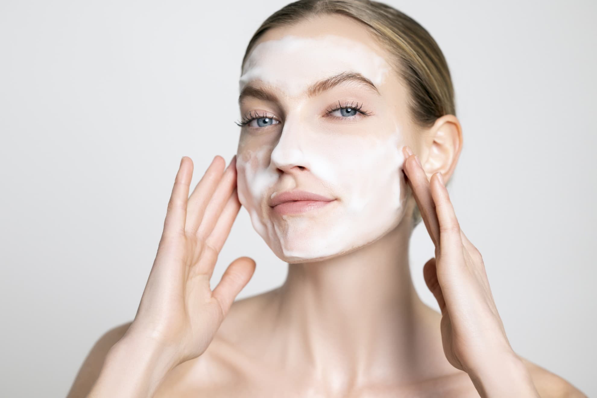 Donna applica mousse detergente sul viso con le dita, viso pulito, pelle luminosa, primo piano su sfondo neutro.
