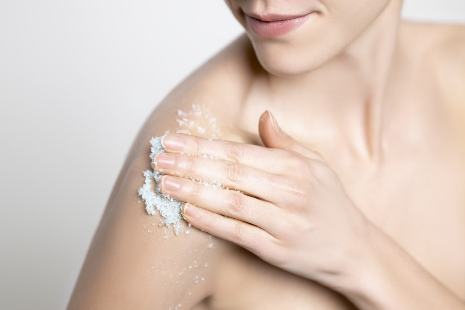 Donna applica lo scrub corpo sul braccio, pelle luminosa, primo piano su sfondo neutro.